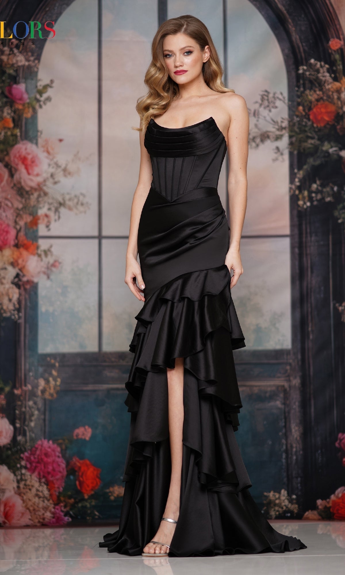 A woman wears the Strapless Long Tiered Prom Gown: Colors Dress 3823, featuring a fitted bodice and ruffled skirt, standing before a floral and window backdrop.